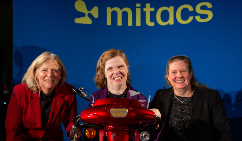 Three women pose for a photo in front of blue background with the middle women in a motorized wheelchair.